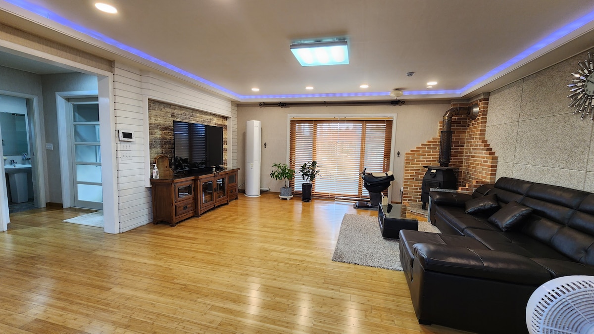 A spacious living area showcases a sleek black sofa opposite a wooden entertainment unit. Natural light filters through a large window adorned with wooden blinds. A small potted plant adds a touch of greenery, while a modern light fixture enhances the inviting space.