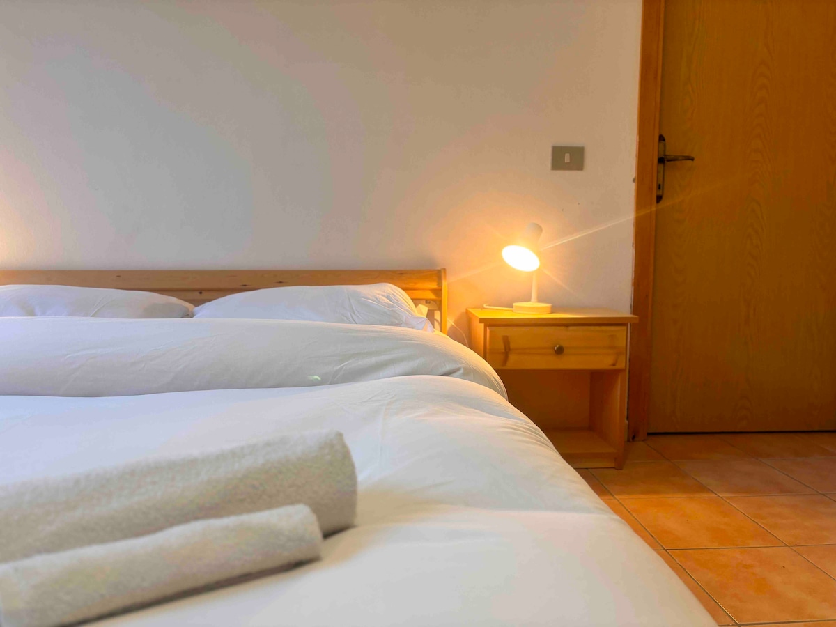 A spacious sleeping area features a light-colored wooden bed with neatly arranged white bedding and rolled towels. A nightstand with a soft-glow lamp adds warmth to the room, positioned next to a wooden door that leads to the hallway.