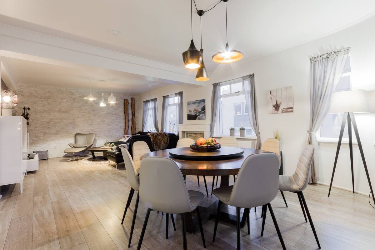A spacious living area features a round dining table surrounded by modern chairs. Elegant pendant lights hang above, illuminating the setting. Light-colored walls and wooden flooring create a warm ambiance, while large windows allow natural light to enhance the contemporary decor.