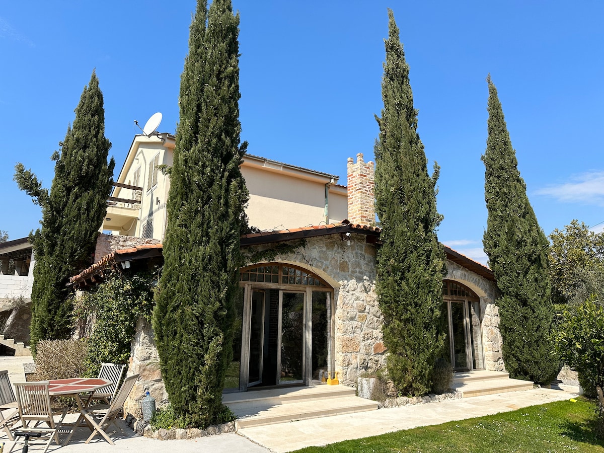 The exterior of a stone house is presented, complemented by tall cypress trees. Large glass doors invite natural light into the living space. A patio area with wooden chairs offers a place for relaxation, surrounded by a well-maintained garden.