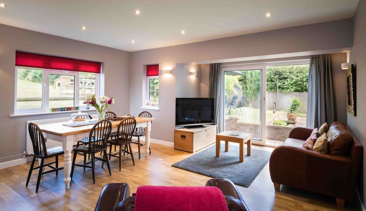 An open plan living and dining area is seen, featuring a sleek television and a comfortable seating arrangement. Large glass doors invite natural light and provide access to the garden. A dining table with chairs is positioned alongside the cozy sofa, creating a welcoming space for relaxation.
