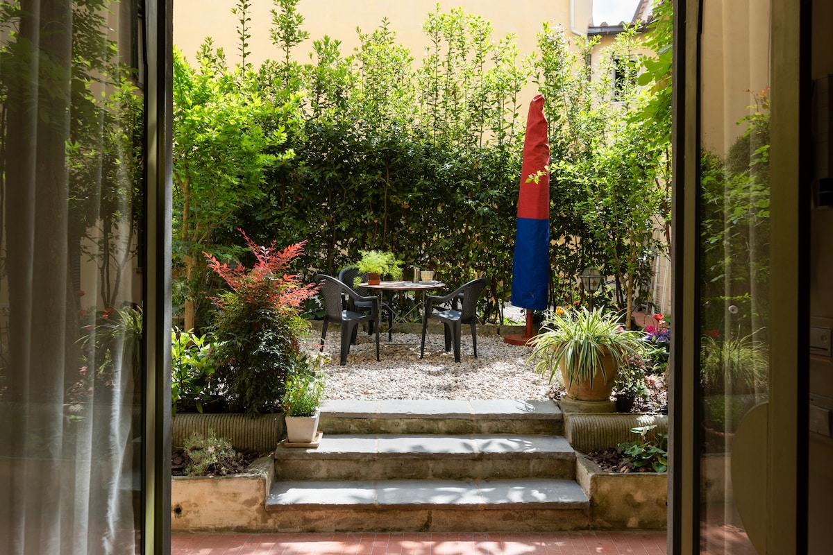 A private garden is visible through the open doors, featuring a gravel area with a table surrounded by four chairs. Lush greenery and colorful plants enhance the space, and a tall umbrella in red and blue provides shade.