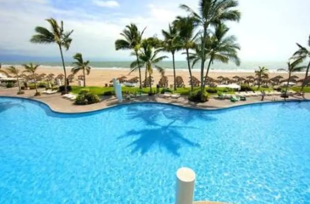 A large, inviting swimming pool is framed by tropical palm trees, with sun loungers positioned along the pool's edge. In the background, the serene beach is visible, leading to the horizon where the sky meets the water.
