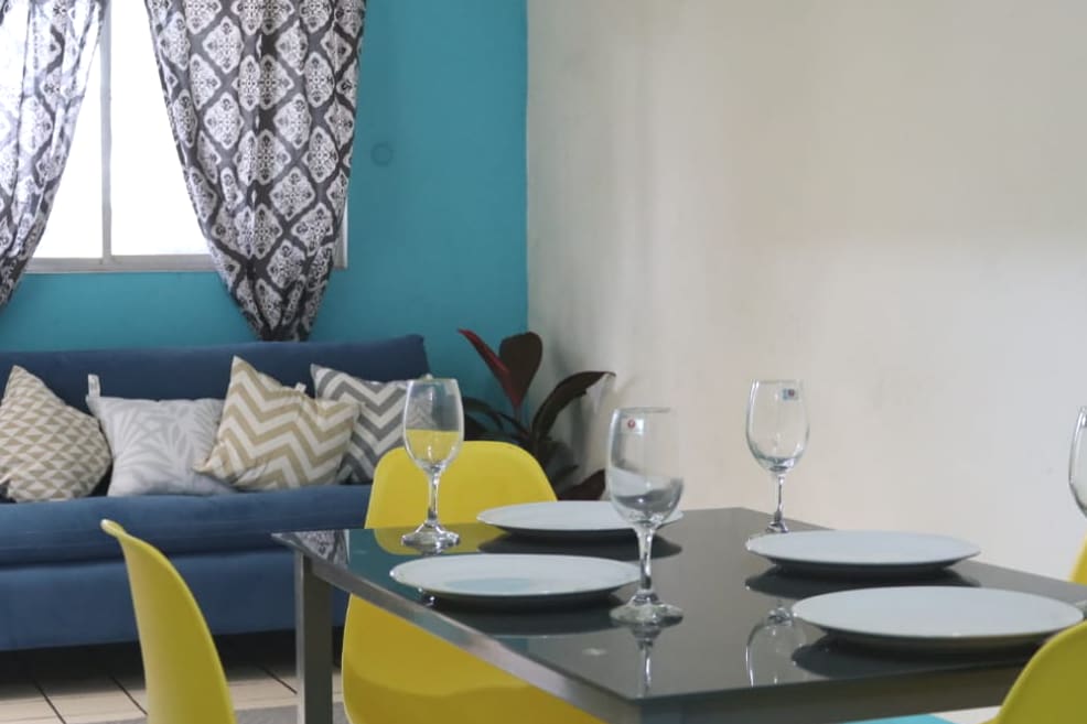 A dining area is displayed with a sleek table set for four, featuring blue plates and clear glassware. A blue sofa with patterned cushions adds comfort, while vibrant yellow chairs provide a cheerful contrast against the light walls.