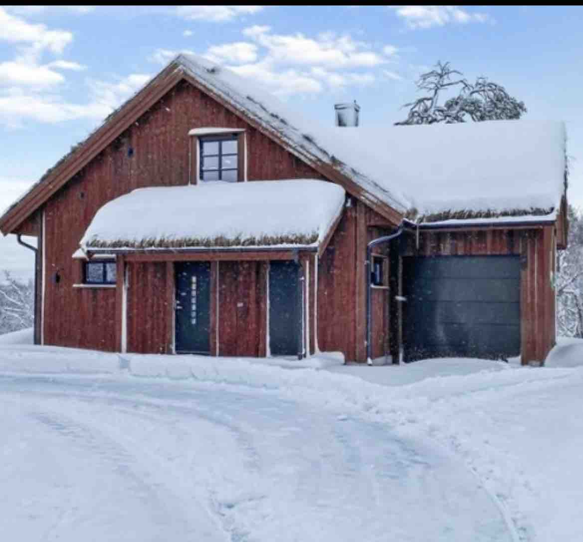 A cozy wooden cabin is surrounded by a blanket of snow, featuring a steeply pitched roof with thatching. The entrance is framed by two dark-colored doors and a large garage door to the side, harmonizing with the tranquil winter landscape.