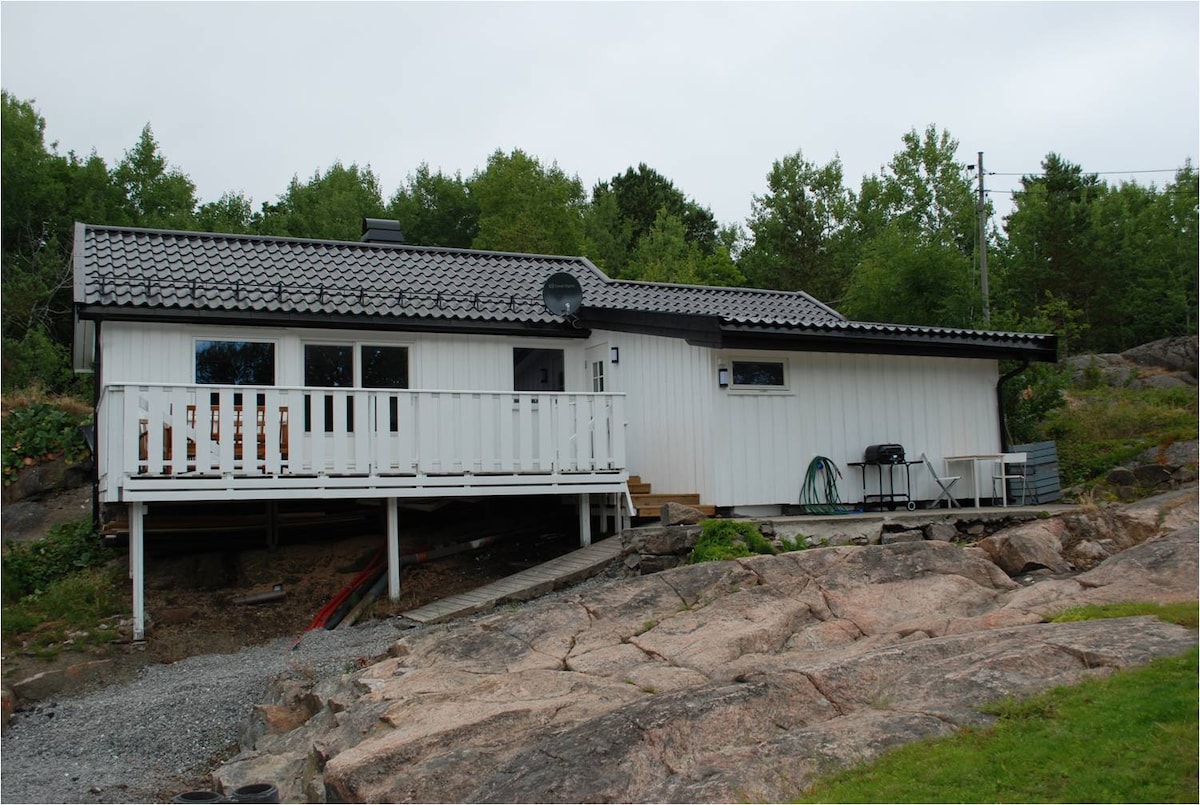 A modern, single-story cabin is positioned on rocky terrain, featuring a light-colored exterior with a dark sloped roof. A deck extends from the front, bordered by white railings. Surrounding greenery complements the natural setting, while gravel pathways provide access to the cabin.