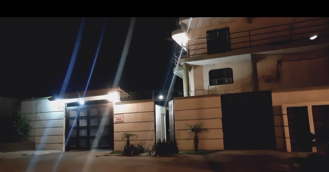 The exterior of a two-story building is shown at night, illuminated by soft lights. A gated entrance is visible, flanked by potted plants. The architecture features a combination of horizontal lines and large windows, giving a modern appearance.
