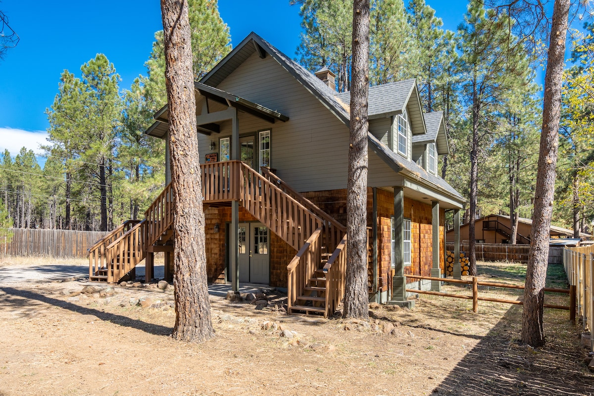 The two-story cottage is surrounded by tall pine trees and features a large front staircase leading to the upper entrance. The exterior is a blend of natural wood and grey siding, complementing the serene forest setting.