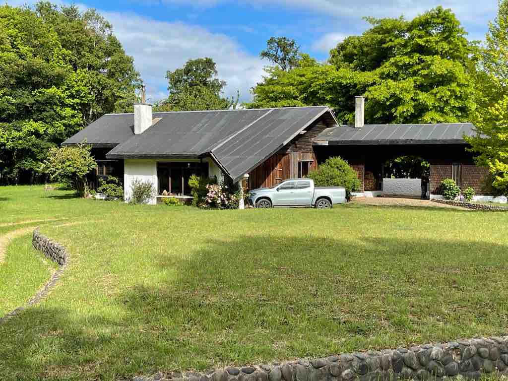 A spacious house constructed from native woods is surrounded by landscaped gardens. The roof features a gentle slope, and a vehicle is parked near the entrance. Lush greenery and trees create a serene natural setting, providing a peaceful atmosphere.