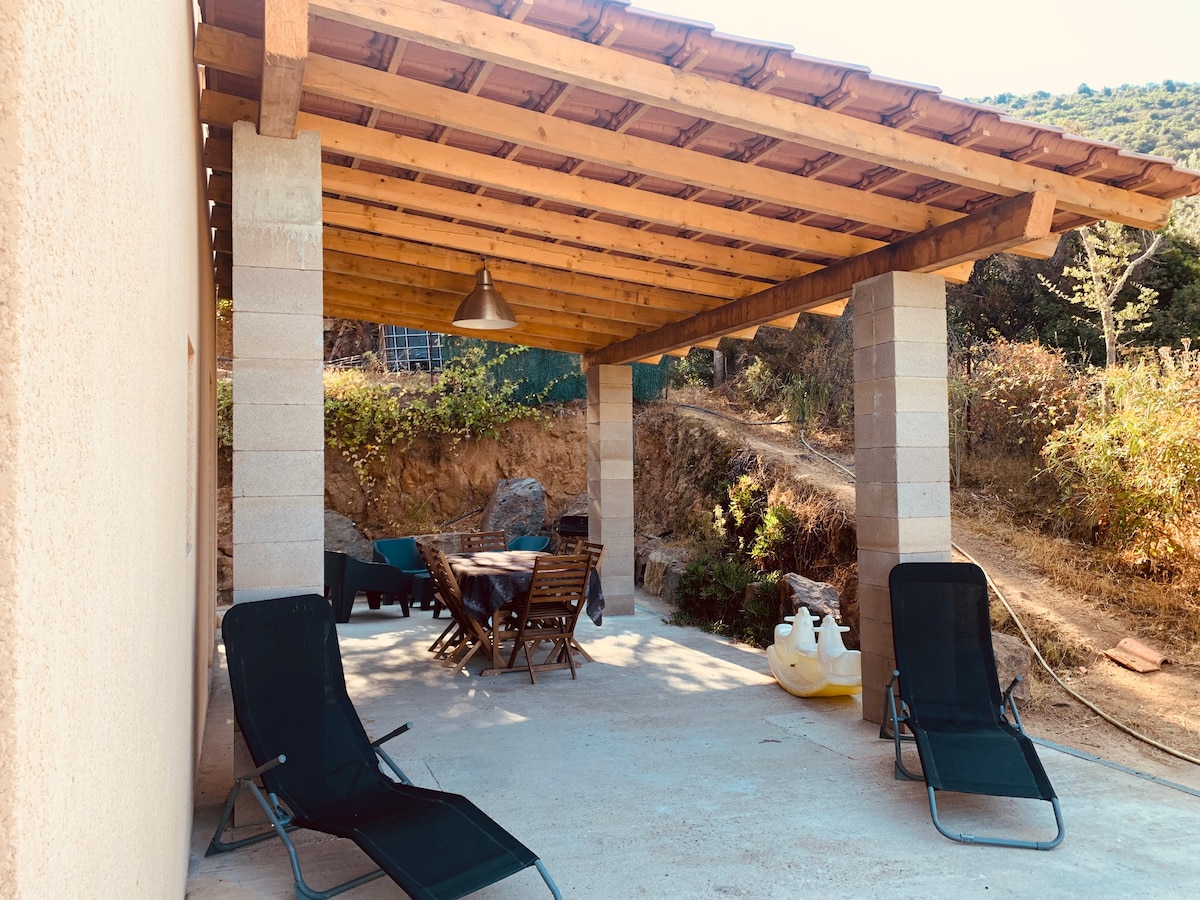 A spacious outdoor terrace is shaded by a wooden overhang, featuring a wooden dining table surrounded by folding chairs. Two loungers are positioned for relaxation. The area is designed to interact with the natural surroundings, with landscaped earth and greenery in the background.
