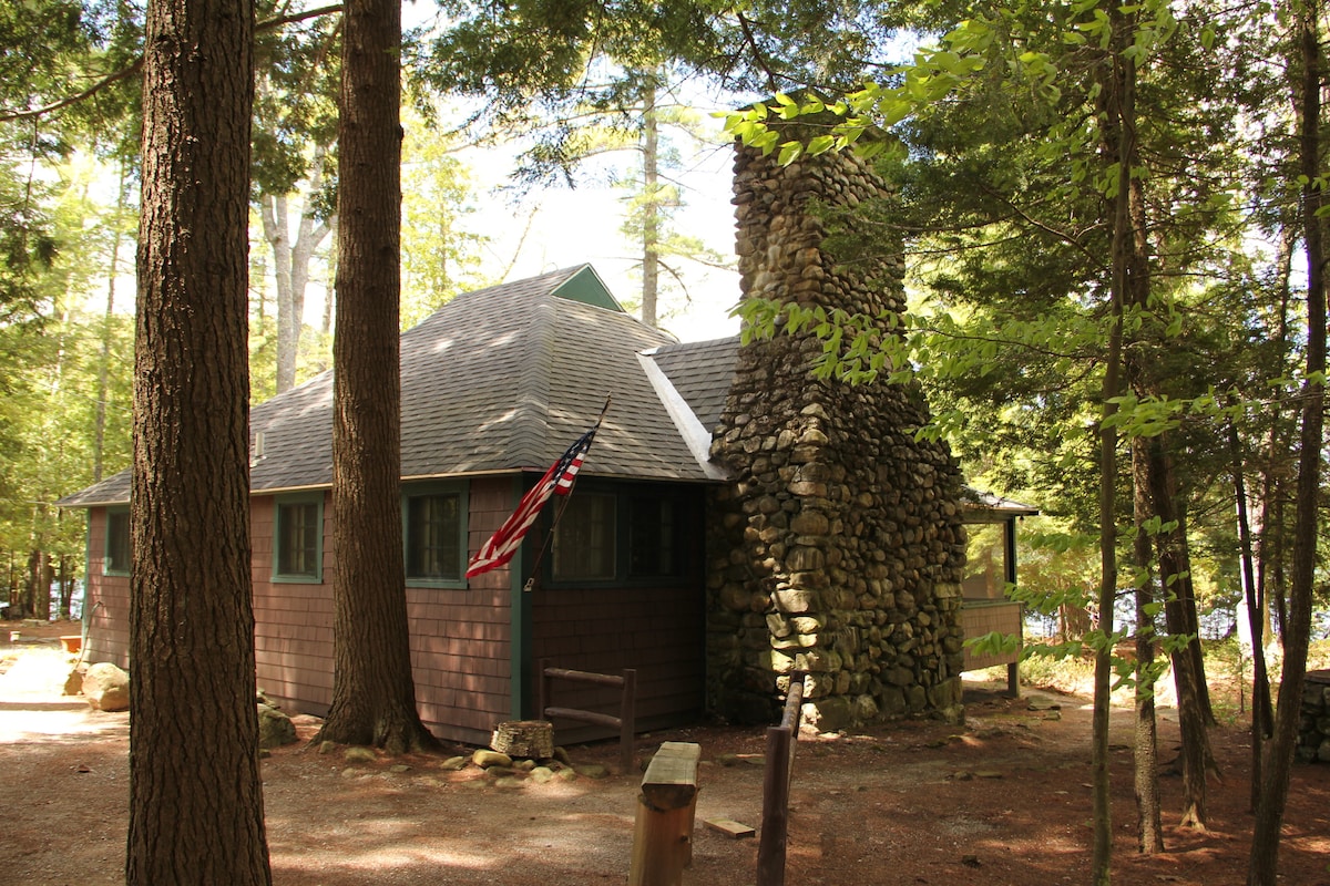 A classic cabin is surrounded by tall trees, featuring a stone chimney and an American flag gently waving. The exterior showcases a combination of dark wood siding and green accents, contributing to a rustic charm, with a pathway leading to the entrance.