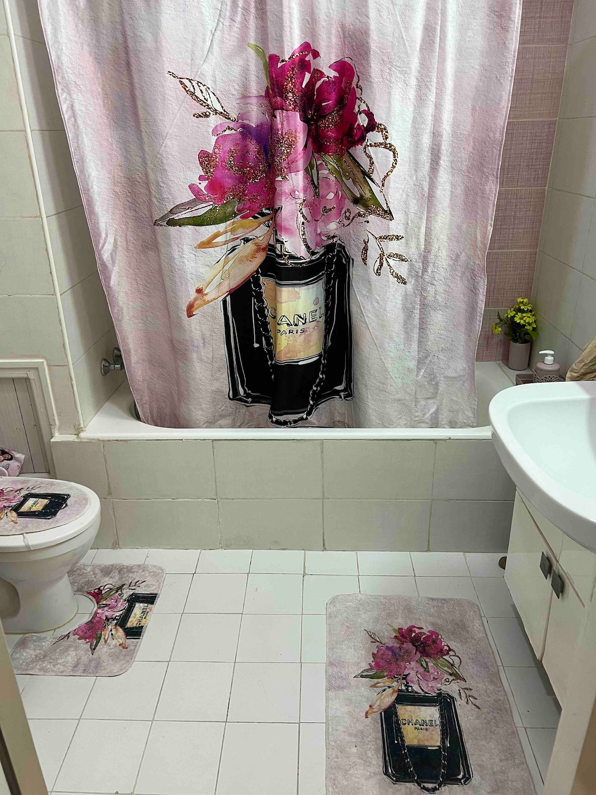 A bathroom is presented with a floral-themed shower curtain featuring a decorative design. The space includes a toilet and a sink, complemented by matching floor mats. A small potted plant adds a touch of greenery, enhancing the overall aesthetic.