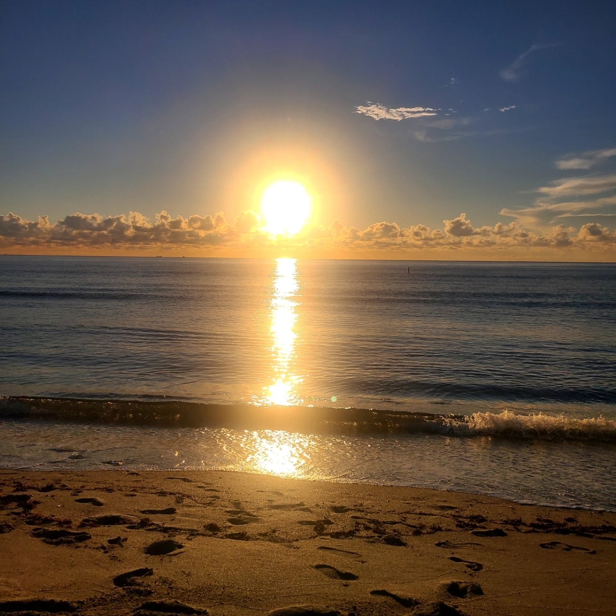 A sunrise casts a golden glow over the water, with sunlight reflecting off the gentle waves. The shoreline is visible in the foreground, featuring soft sand and footprints, while a clear blue sky filled with scattered clouds completes the serene beach setting.
