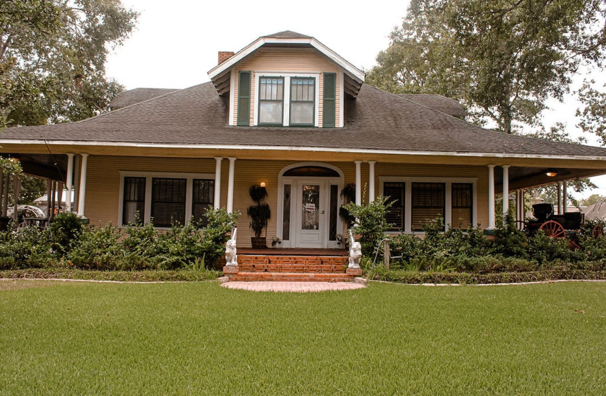 A charming, two-story house features a large front porch supported by white columns and is flanked by neatly maintained flower beds. A well-kept lawn surrounds the home, while lush greenery provides a natural border.