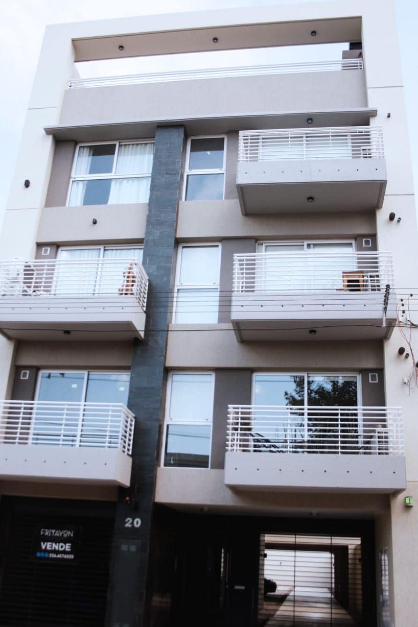 A modern, multi-story building with a contemporary facade is displayed. The structure features multiple balconies with white railings, large windows, and a sleek entrance that leads to the interior. The neutral color palette contributes to a clean and inviting appearance.