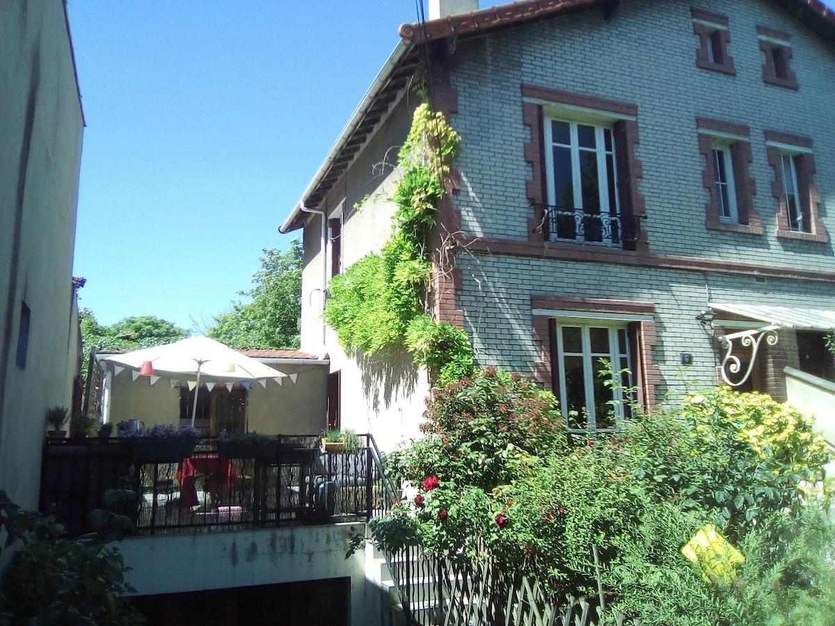 A charming house is presented, featuring a lush garden and a spacious terrace adorned with a large umbrella. Vibrant greenery climbs the exterior walls, while large windows invite natural light into the interior. The peaceful setting is complemented by colorful flowers surrounding the patio.