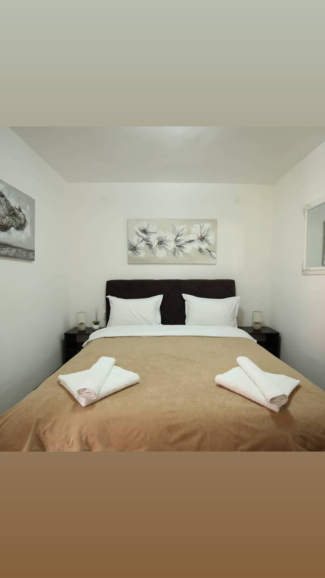 A cozy sleeping area is presented with a spacious bed adorned with light-colored linens. Two neatly folded towels are placed at the foot of the bed. A floral artwork and a mirror are mounted on the walls, enhancing the soft and neutral color palette.