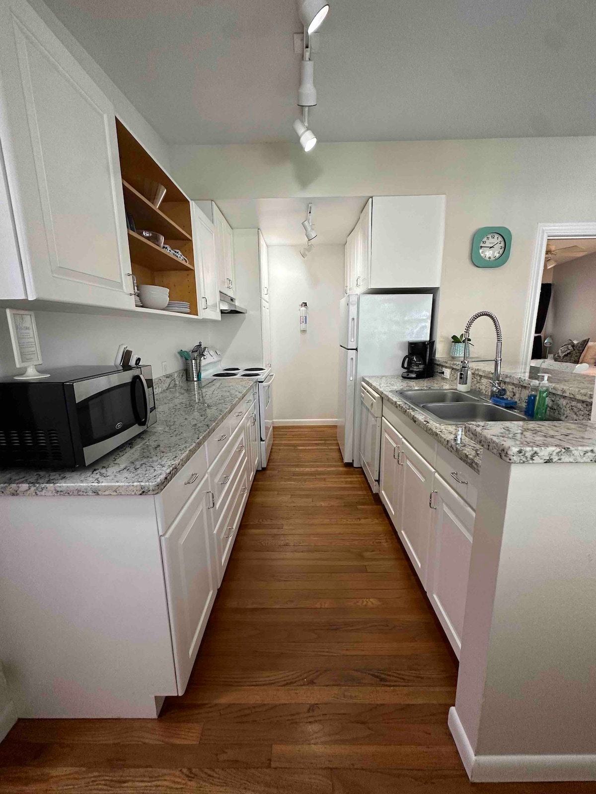 A modern kitchen is featured, with white cabinetry and granite countertops. Appliances include a microwave, stove, refrigerator, and coffee maker. Ample storage is provided through open and closed shelving, allowing for easy access to kitchenware. Warm wood flooring complements the design.