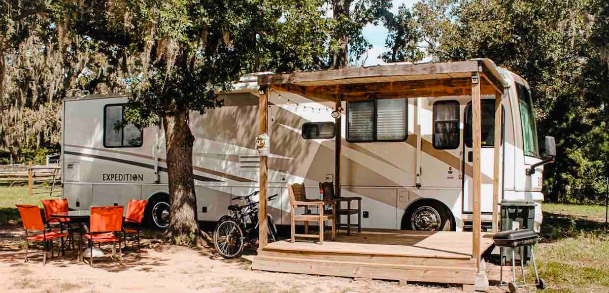 A spacious motorhome is parked under a tree, featuring a wooden deck with seating and a table. Four outdoor chairs complement the setup, while a bicycle rests nearby. The surrounding area showcases a grassy landscape typical of a farm setting.