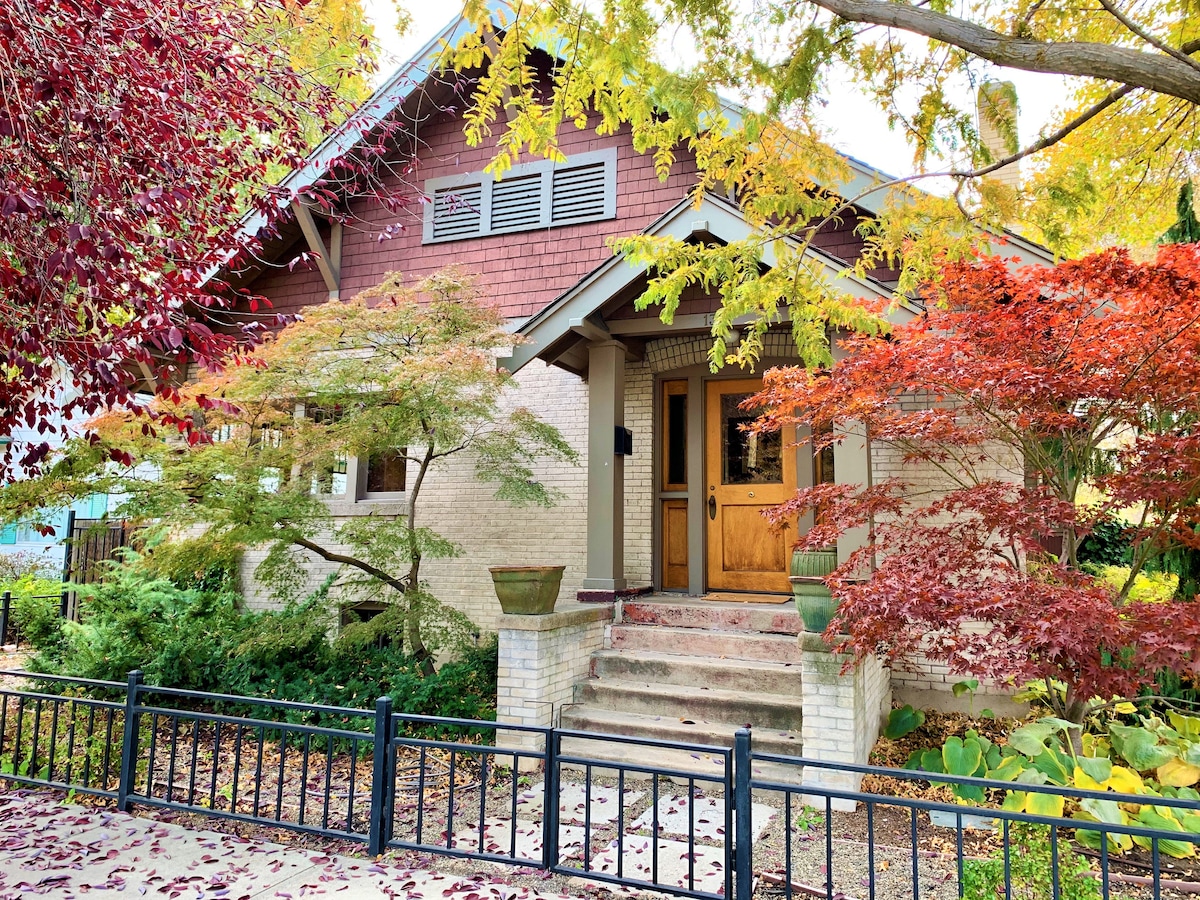 The home features a charming entry with a wooden door surrounded by ornate sidelights. Seasonal foliage in shades of red and orange enhances the welcoming atmosphere. Steps lead up to the entrance, bordered by greenery and decorative planters.