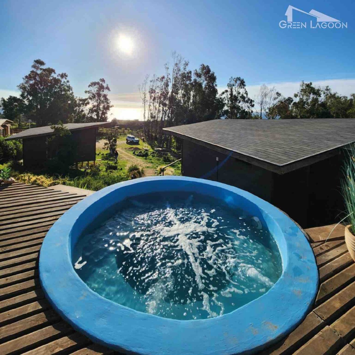 A circular blue jacuzzi is set on a wooden deck, surrounded by trees and a view of the ocean in the background. The sun shines brightly above, creating a serene atmosphere. Nearby structures are visible, blending into the natural landscape.