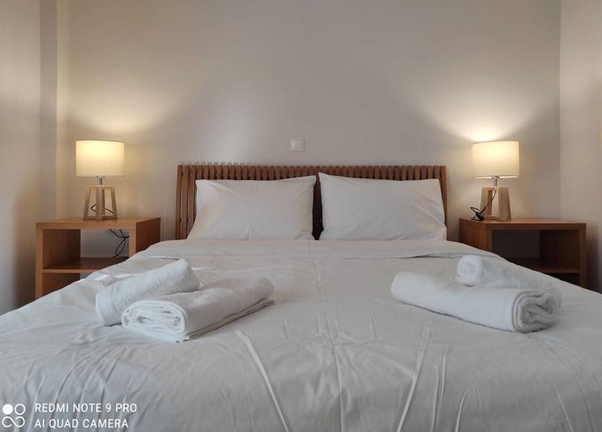 A modern bedroom features a spacious bed with crisp white linens. Nightstands with warm lamps are placed on either side. Neatly folded towels are arranged at the foot of the bed, enhancing the minimalist design.