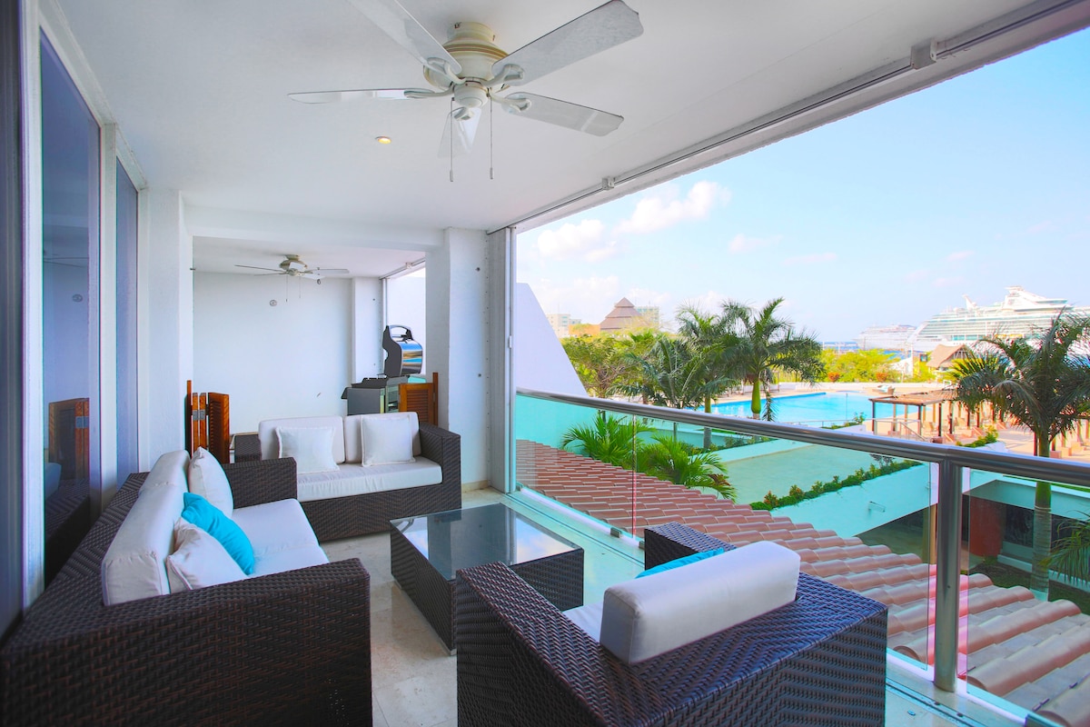 A private balcony features a comfortable seating area with wicker furniture and plush cushions. A glass table anchors the space, with stunning views of the ocean and pool visible through expansive glass doors. Ceiling fans provide airflow in this relaxing outdoor setting.
