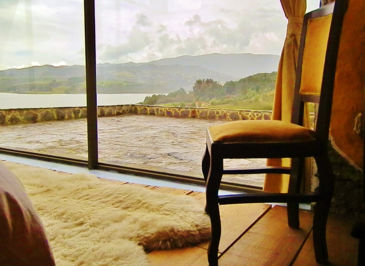 A wooden chair is positioned beside a large glass window, offering a view of a serene landscape with hills and water. A soft rug lies on the floor, adding texture to the space. Natural light fills the room, creating a calm atmosphere.