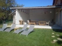 A covered terrace area is visible, featuring a long wooden table and several chairs. Four lounge chairs are arranged on the grassy lawn, providing comfortable seating. The space is well-lit by natural sunlight, and a serene outdoor setting is highlighted by the surrounding greenery.