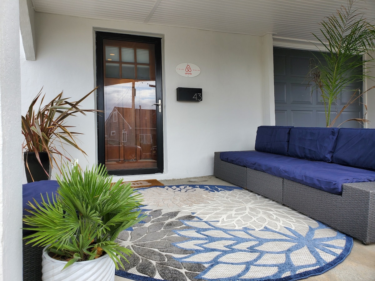 An inviting outdoor seating area features a dark wicker sectional sofa and a round, patterned area rug. Potted plants add a touch of greenery. The entrance is marked by a black door with a window, and a sign indicates the listing number.