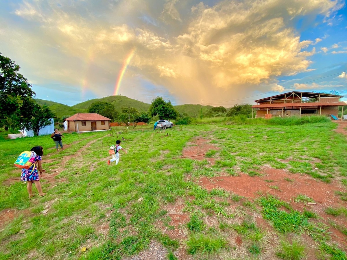 An expansive grassy area is visible, featuring a small cabin and a larger structure in the background. A rainbow arches over the scene, while individuals can be seen walking and enjoying the outdoors. Lush greenery adds a vibrant touch to the landscape.