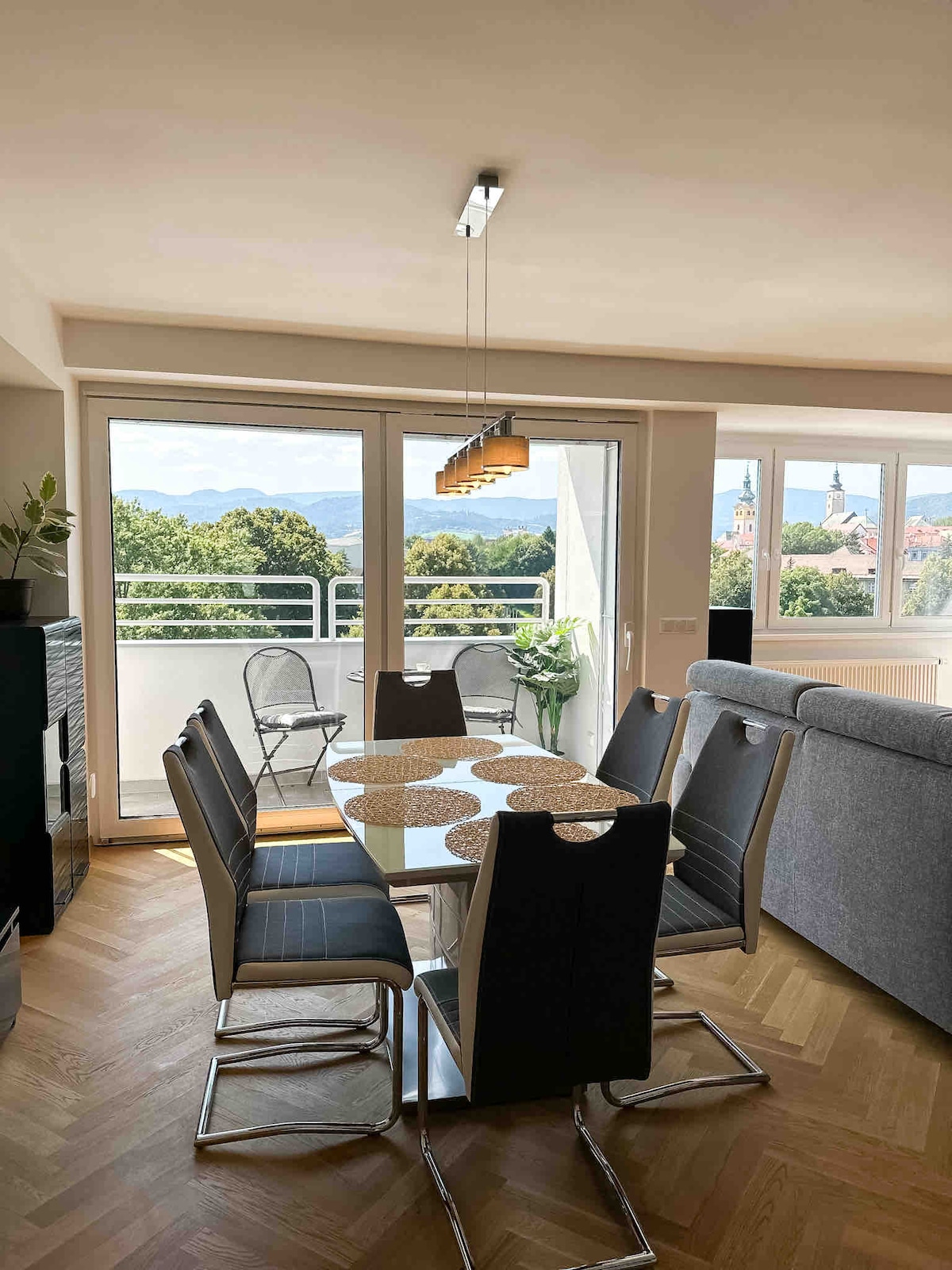 A modern dining area features a round glass table surrounded by six sleek chairs. A scenic view of mountains and greenery is visible through large windows, with a pair of chairs positioned on the balcony. A contemporary light fixture hangs above the table.
