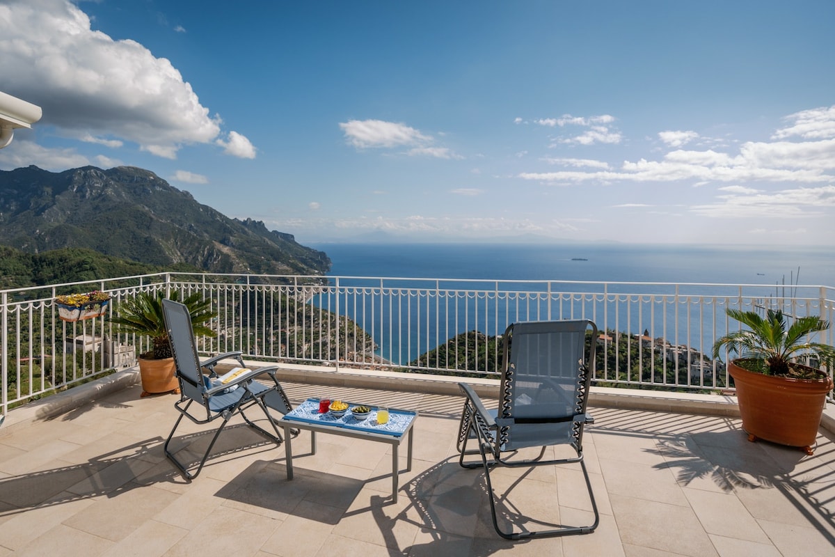 A spacious terrace offers scenic views of the coastline and mountains. Two lounge chairs are positioned beside a small table, which displays colorful items. Potted plants add a touch of greenery, enhancing the serene outdoor setting.
