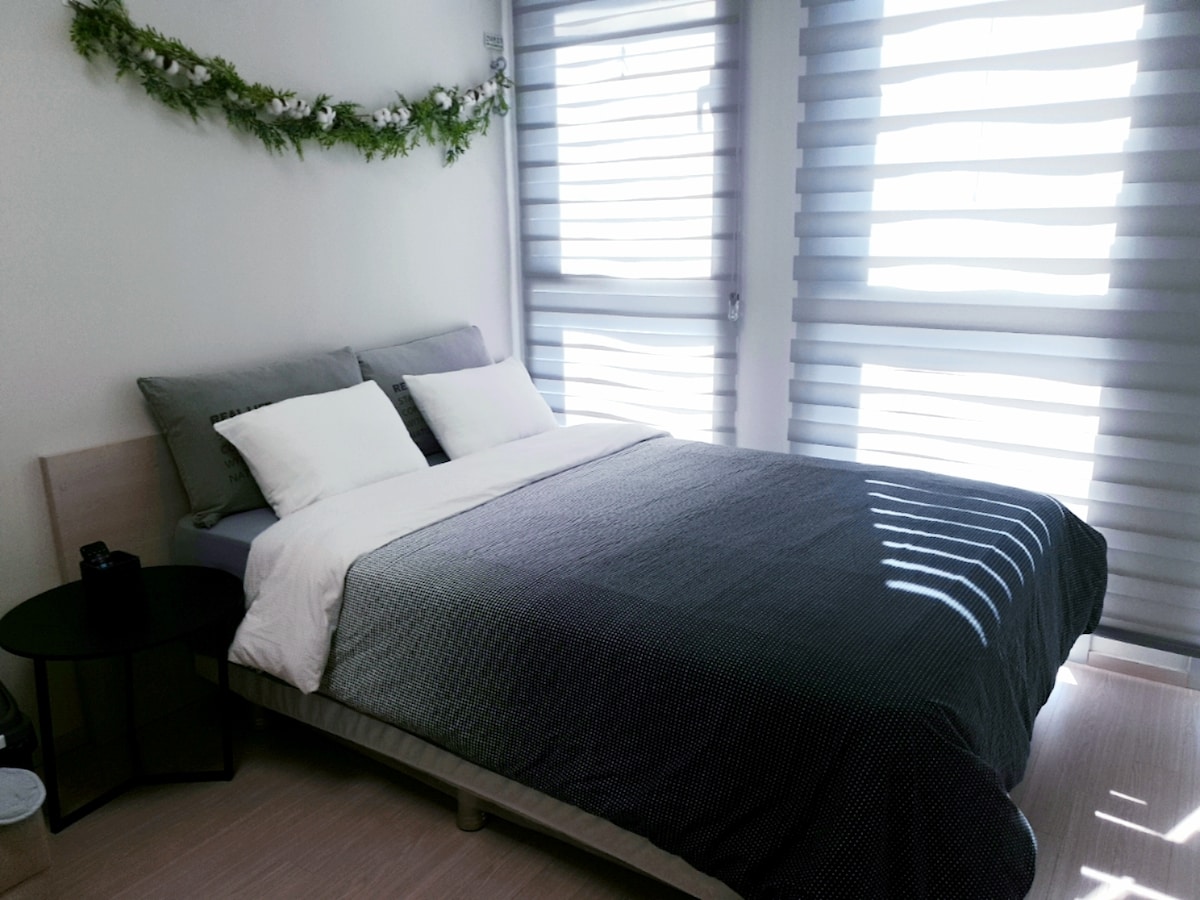 A bright sleeping area features a spacious bed dressed in neutral tones. Large windows with light-filtering shades provide natural light and a glimpse of greenery. A small bedside table stands beside the bed, complementing the modern decor.