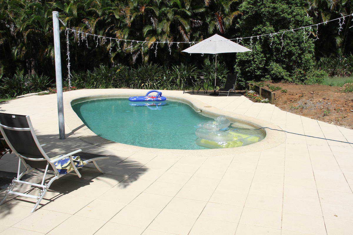 A private swimming pool features a unique shape, surrounded by a spacious patio. Lounge chairs provide seating, while a shaded area with an umbrella offers a cool retreat. Inflatable floats rest on the water, and lush greenery borders the scene, enhancing the tranquil atmosphere.