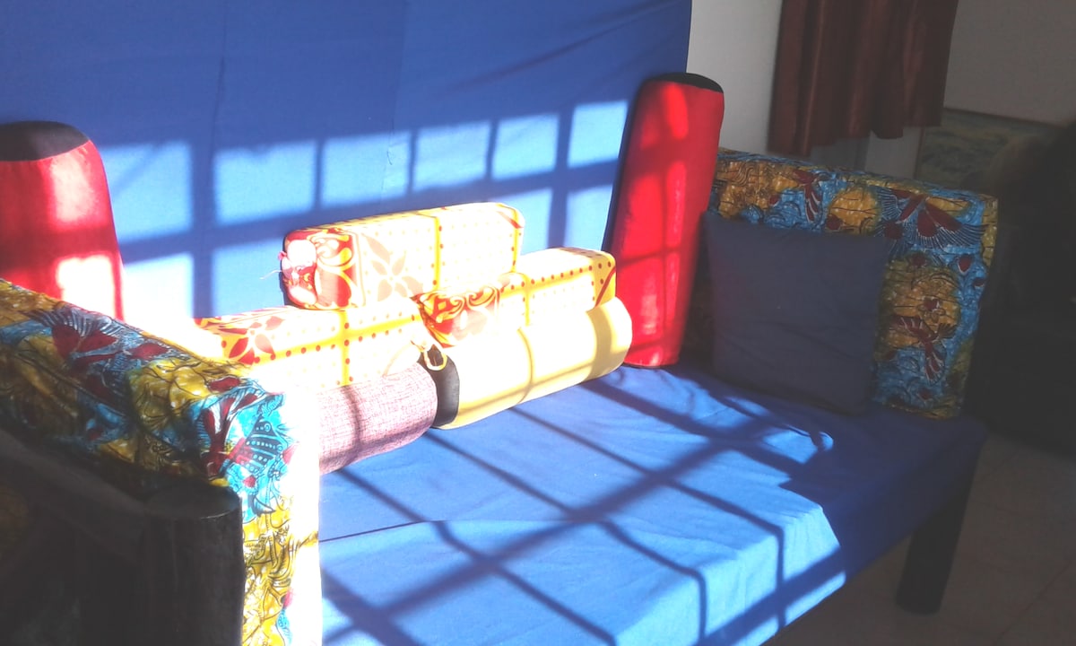 A colorful seating area is presented with a blue backdrop, featuring a sofa adorned with bright fabric patterns. Cushions and rolled pillows in coordinating colors are arranged neatly, while sunlight casts a decorative shadow across the space.