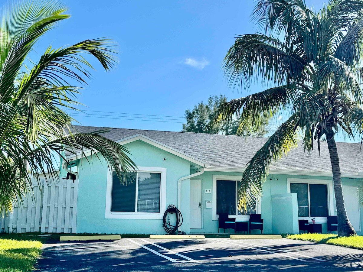 The exterior of a light blue bungalow is framed by palm trees and a well-maintained lawn. A paved parking area is visible in front, along with a porch featuring two chairs and a decorative wreath by the door.