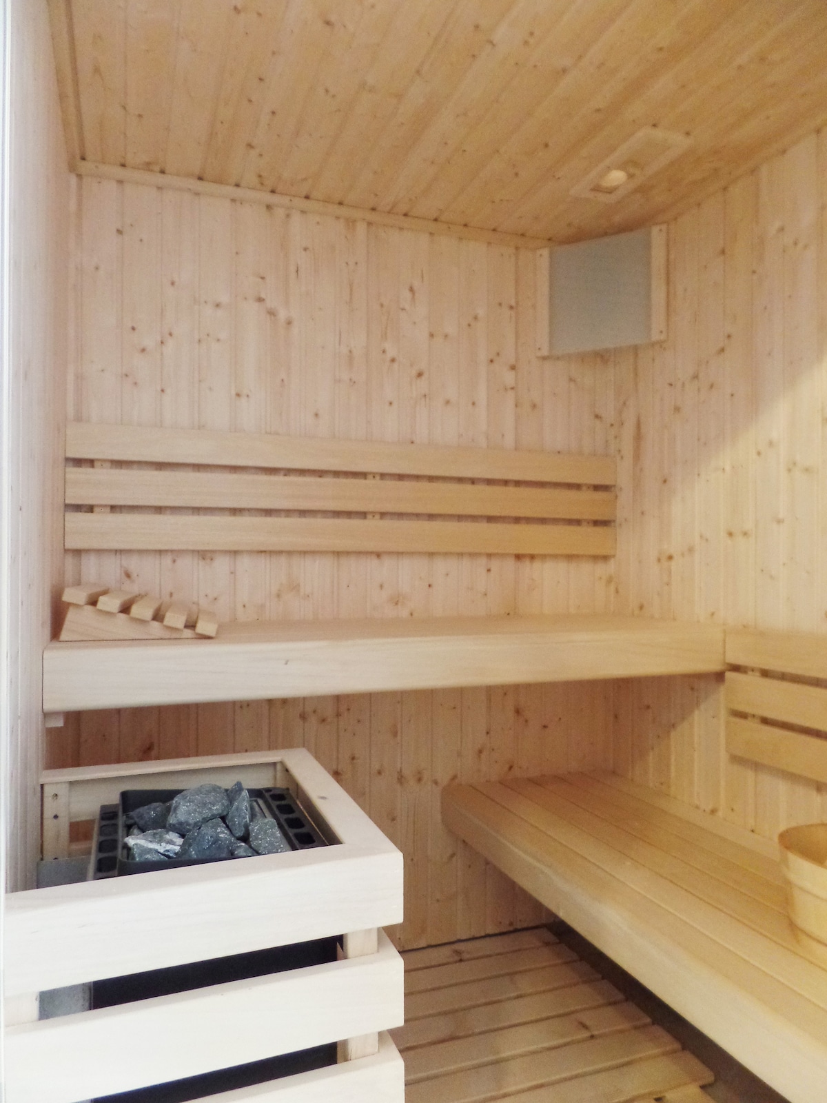 A sauna interior is shown with natural wood paneling. Two benches are positioned along the walls, providing seating for relaxation. A stone-filled heater is visible, contributing to the sauna's warm atmosphere. The simple design enhances the calming environment, ideal for unwinding.