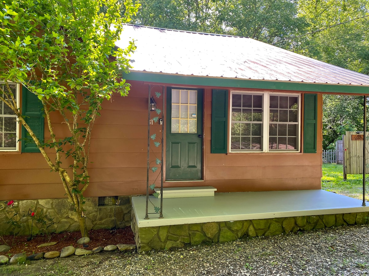 A charming cabin exterior is showcased with a welcoming green door. Multiple windows provide natural light, framed by dark green shutters. A small porch area with a concrete surface is bordered by a stone foundation and lush greenery, enhancing the rustic appeal.