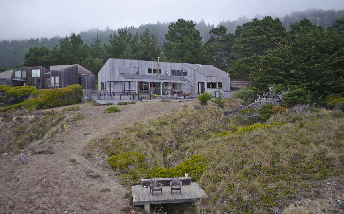 A modern home with a sleek design is positioned on a coastal bluff, surrounded by natural greenery. The property features a spacious deck with outdoor seating, providing views of the distant ocean and surrounding landscape under a soft, overcast sky.