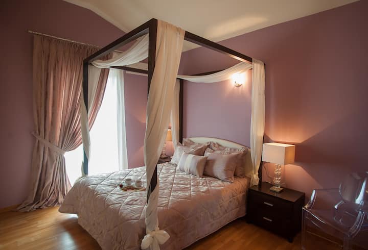 Chumba cha kulala cha watu wawili chenye kitanda cha nguzo nne na mashuka yaliyopambwa na mapazia mazito ambayo huunda mandhari ya kifalme.