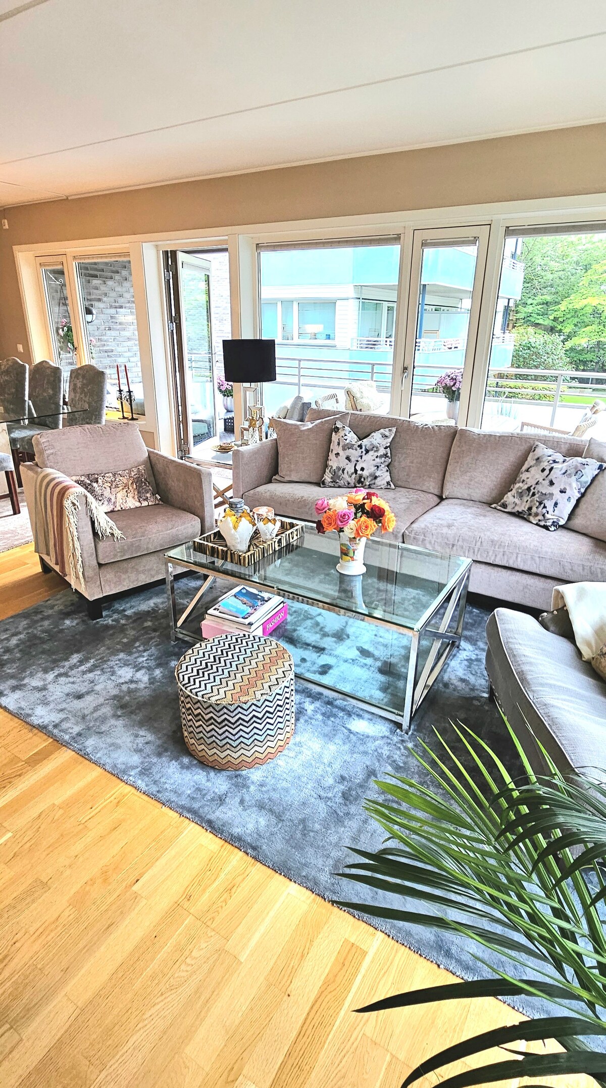 An inviting living room features a comfortable sofa set, a glass coffee table, and decorative accents. Large windows allow ample natural light, offering views of the greenery outside. A patterned rug adds texture to the wooden floor, enhancing the space's relaxed ambiance.