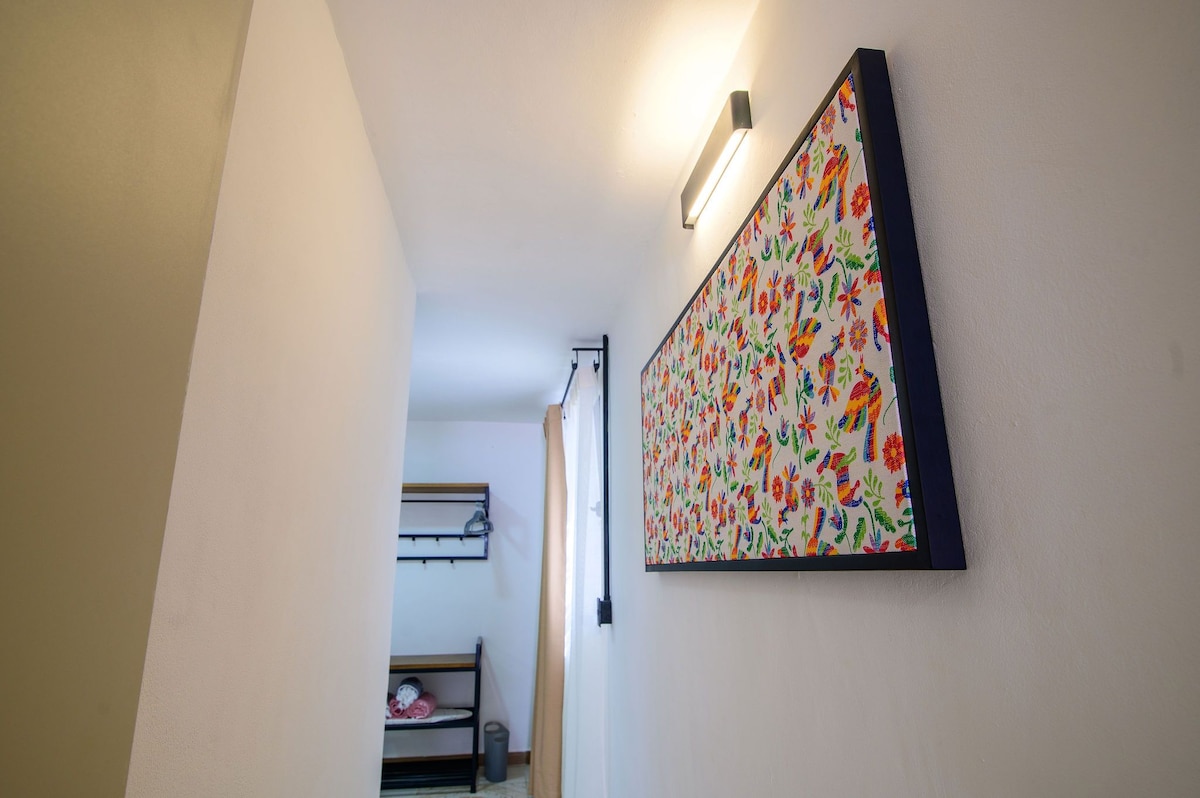 A brightly lit hallway is lined with a colorful floral artwork, adding vibrancy to the space. To the side, a wooden rack offers hanging space, while a bunk bed is visible in the background. Soft natural light filters in from a window.