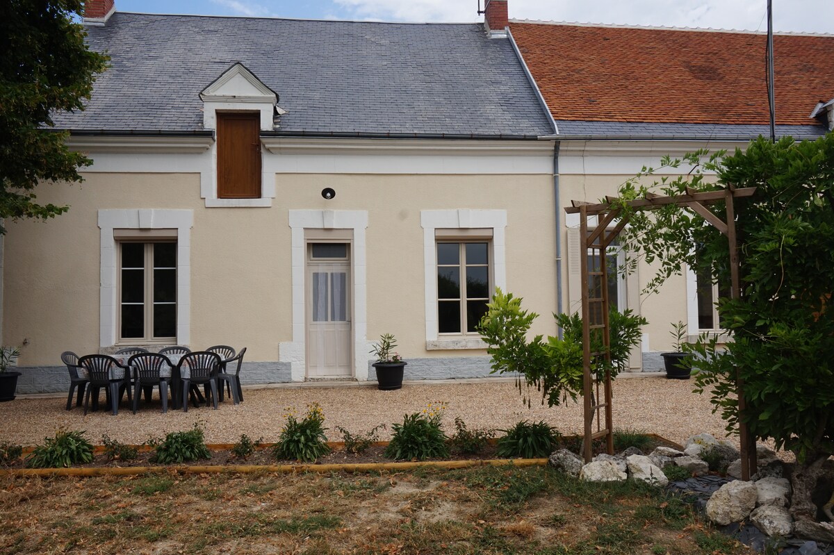 The front of the house showcases a light-colored façade with a symmetrical arrangement of windows. A wooden door is centrally positioned. A gravel area features a table and several chairs. Lush greenery surrounds the entrance, contributing to a serene outdoor setting.