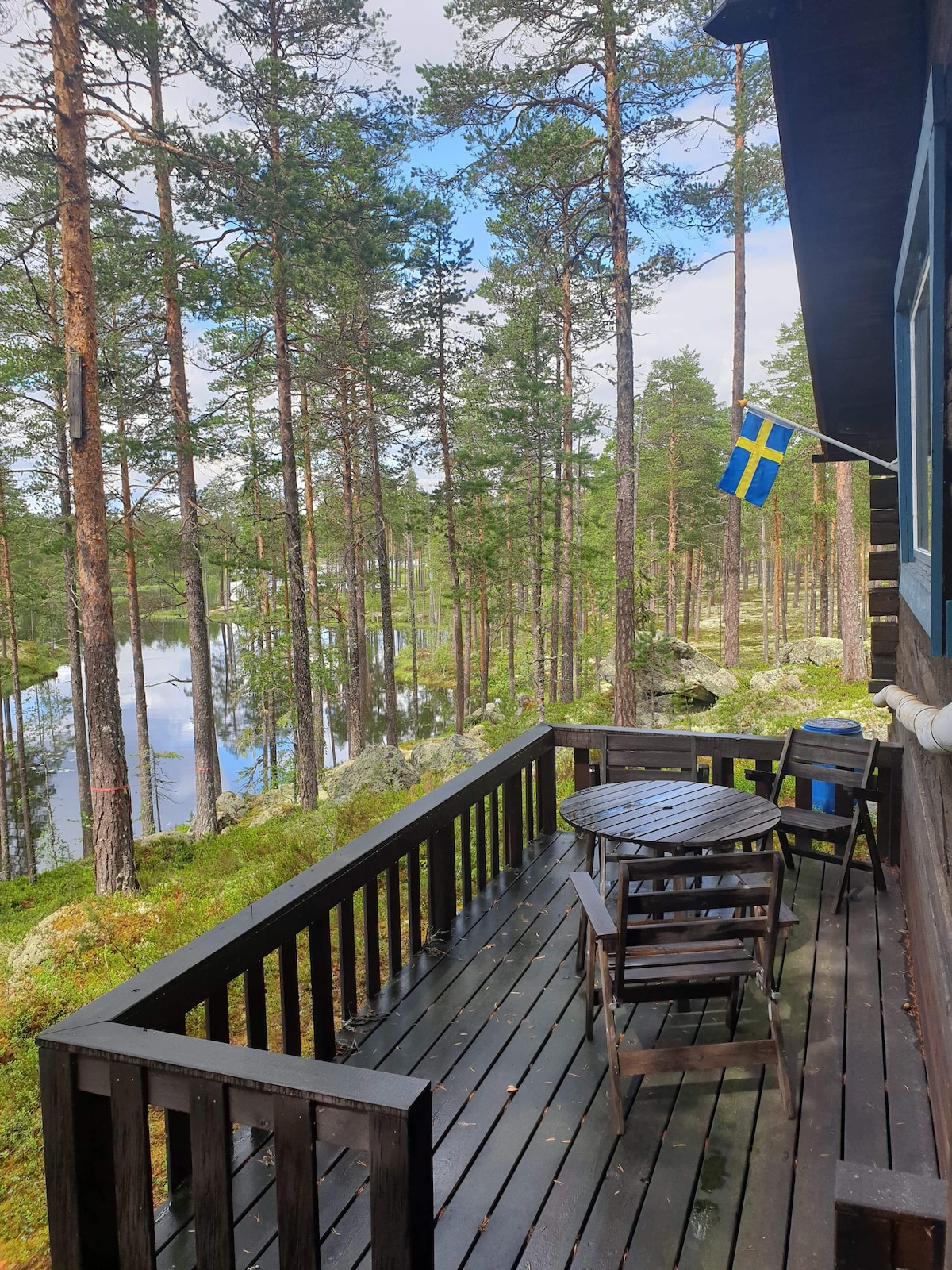A wooden deck is visible, featuring a round table and two chairs. A Swedish flag is displayed on the side. The surrounding area includes tall trees and a reflective body of water, creating a serene forest setting.