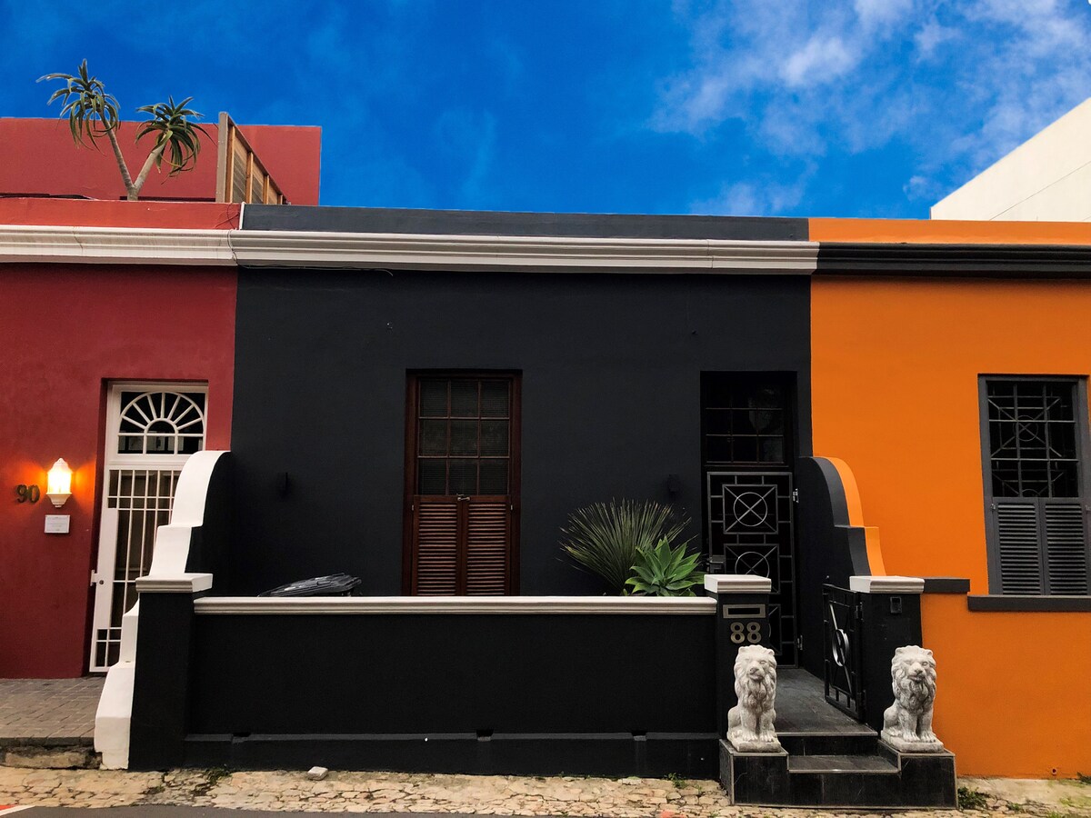 A vibrant street scene showcases a charming building facade in contrasting colors. The black structure features a decorative entrance with classic ironwork and lion statues flanking the front steps. Nearby buildings display hues of red and orange, framed by a clear blue sky.