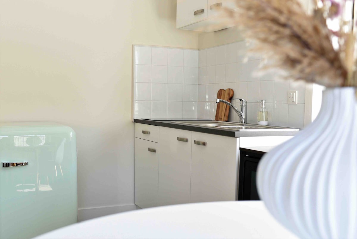 A modern kitchenette features sleek white cabinetry and a stainless-steel sink. A small green refrigerator stands nearby, complemented by wooden cutting boards displayed on the counter. Decorative items, such as a vase with dried flowers, add a touch of warmth to the space.