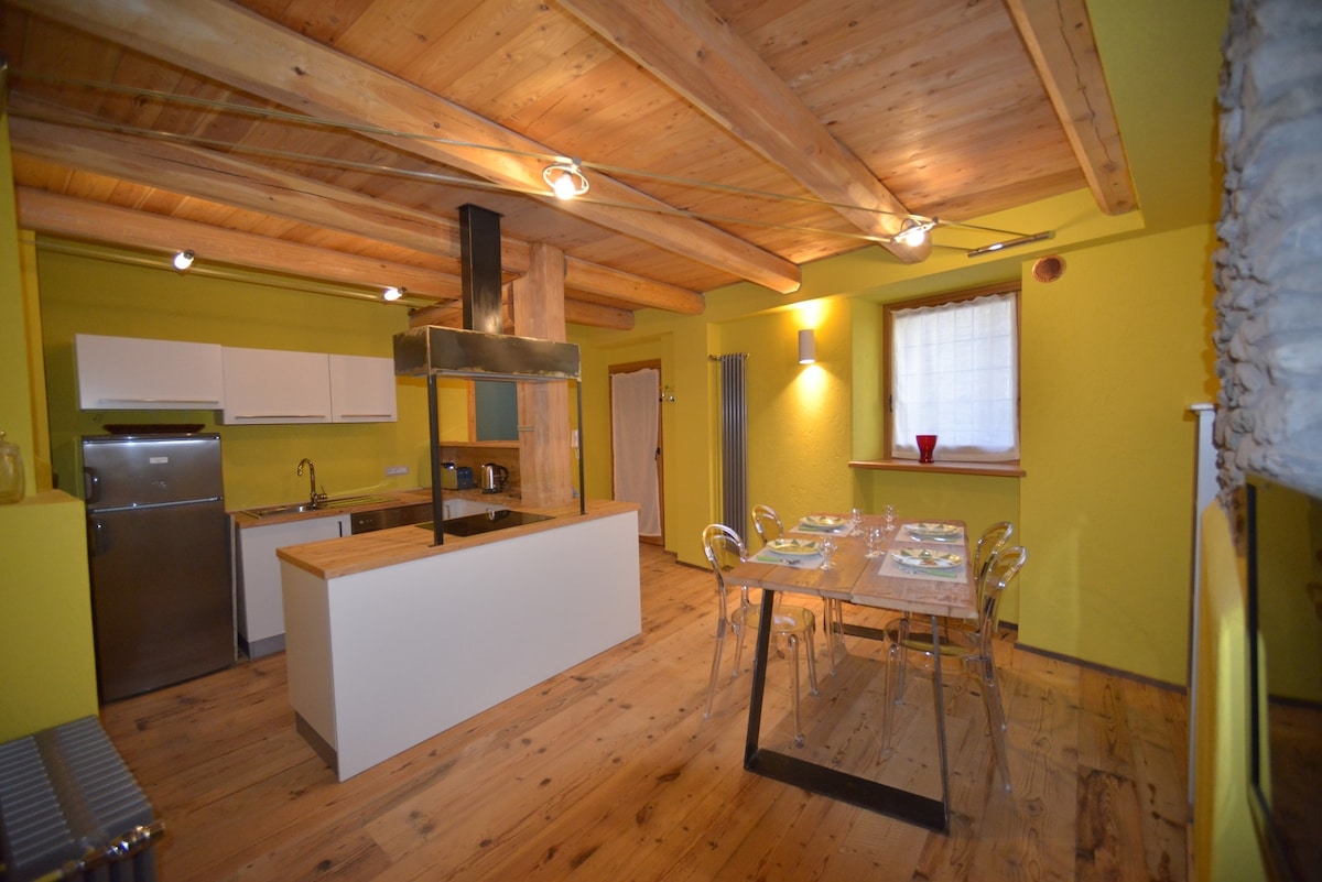 An open-plan kitchen and dining area features wooden beams and bright yellow walls. A stainless steel refrigerator and modern appliances are visible, complemented by a wooden dining table set for four. Natural light filters through a window with simple curtains, enhancing the welcoming atmosphere.