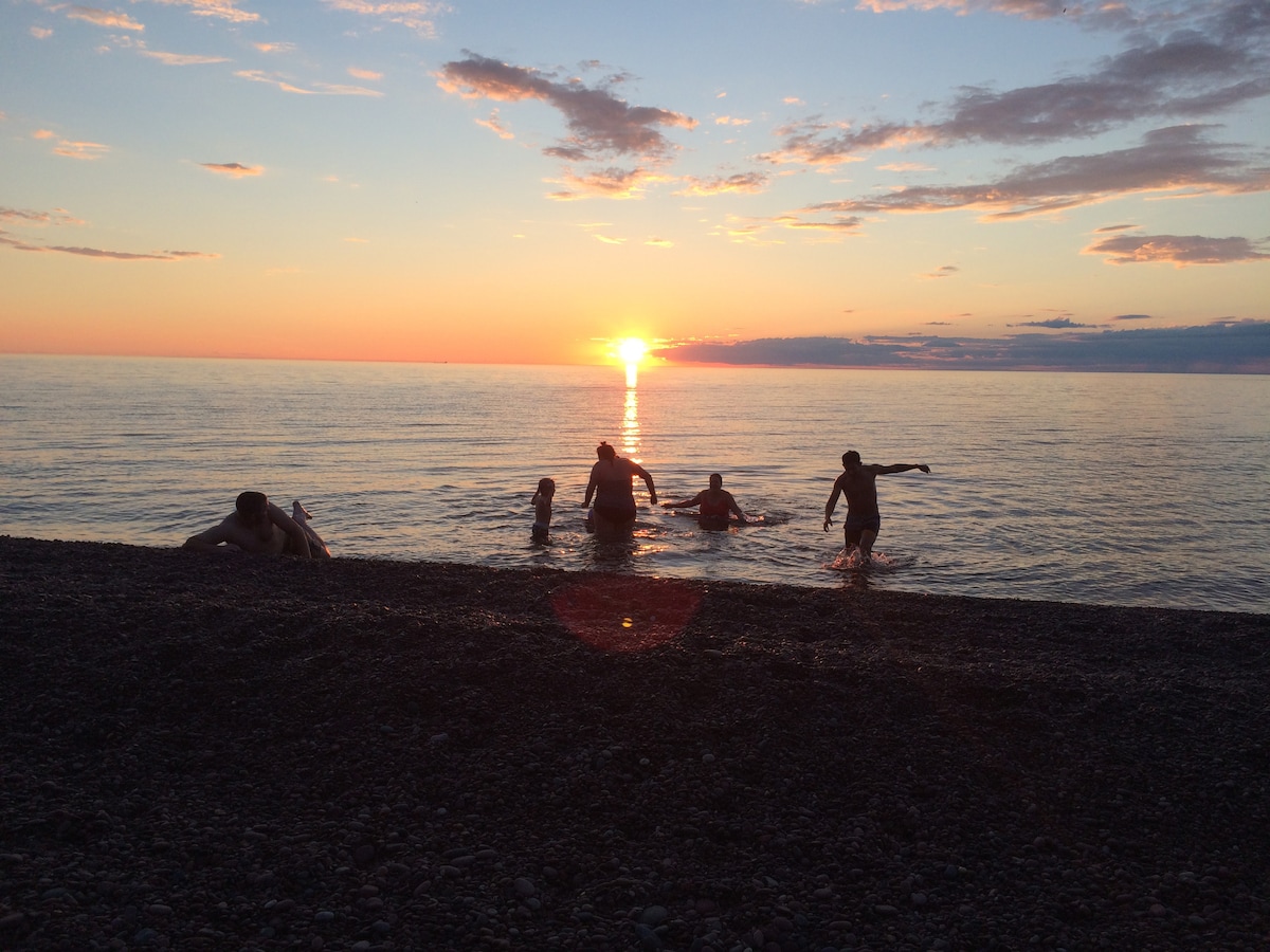 A serene beach scene showcases individuals enjoying a swim in calm waters as the sun sets on the horizon. Shades of orange and pink fill the sky, reflecting on the surface of Lake Superior, while the shoreline features smooth pebbles beneath their feet.