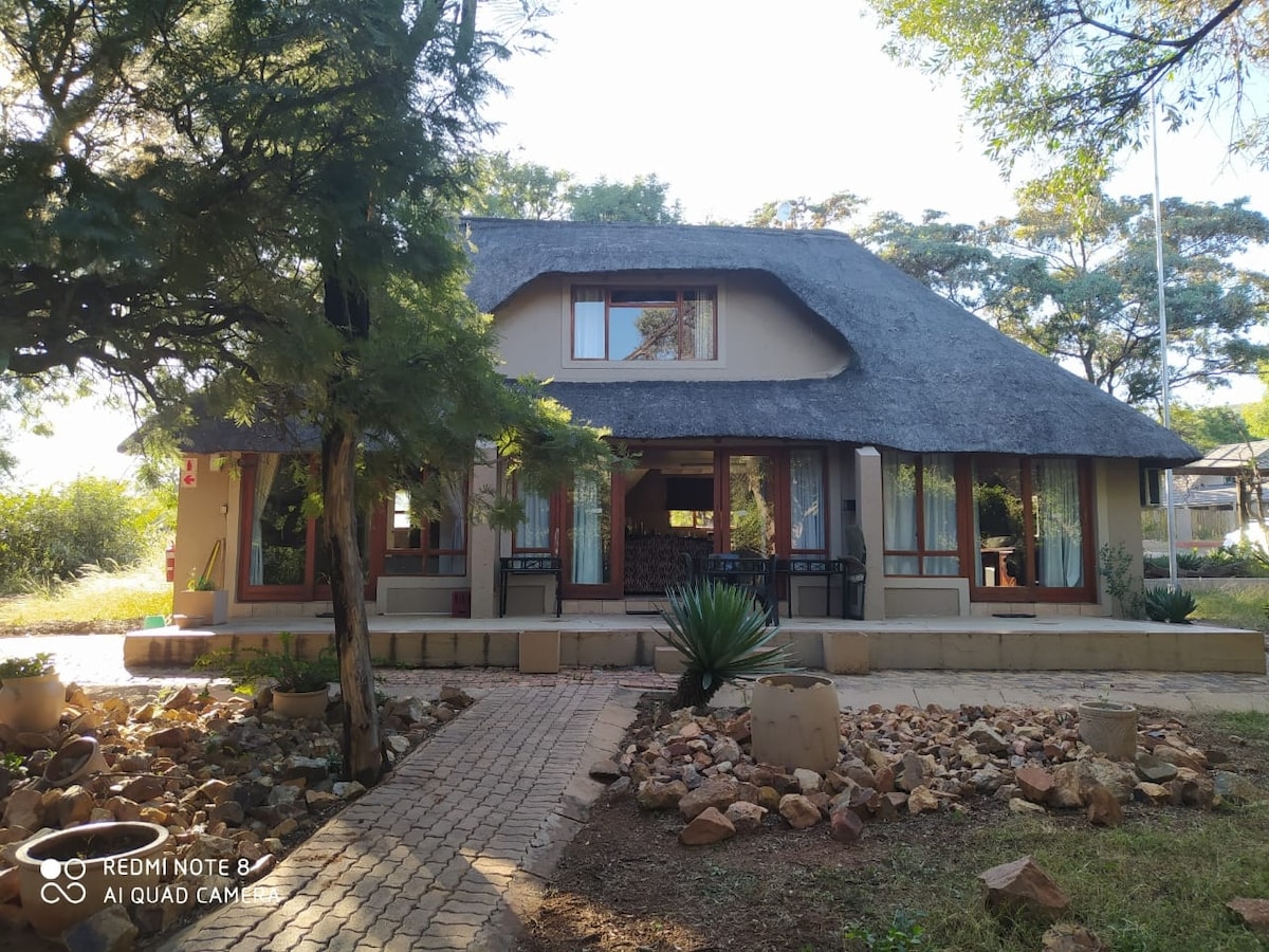 The exterior of a charming chalet with a thatched roof is presented, surrounded by lush greenery. A stone walkway leads to a covered porch, where outdoor seating is visible. The building showcases large windows that offer views into the inviting interior.